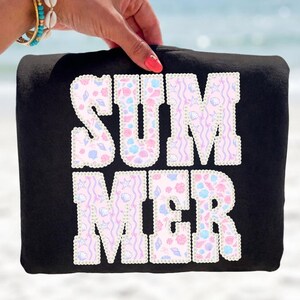 May include: Black tote bag with the word "SUMMER" in white, outlined with pearls. The letters are filled with a pastel seashell pattern. The bag is held up against a blurred beach background.