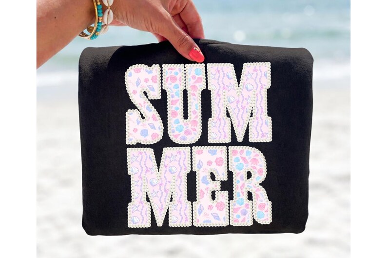 May include: Black tote bag with the word "SUMMER" in white, outlined with pearls. The letters are filled with a pastel seashell pattern. The bag is held up against a blurred beach background.