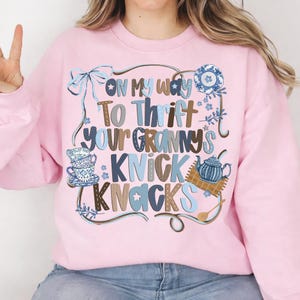 May include: A light pink sweatshirt with the phrase "ON MY WAY TO THRIFT YOUR GRANNY'S KNICK KNACKS" in a mix of blue and brown fonts. The design includes illustrations of teacups, a teapot, and floral accents.