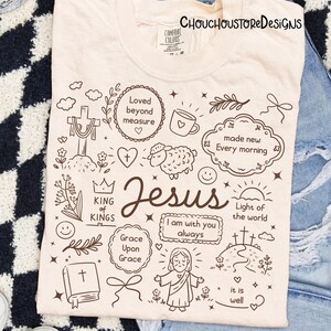 May include: Cream-colored t-shirt with a hand-drawn design featuring the word "Jesus" and various religious symbols and phrases, including a cross, heart, and "Light of the world". The text "CHOUCHOUSTORE DESIGNS" is at the top.