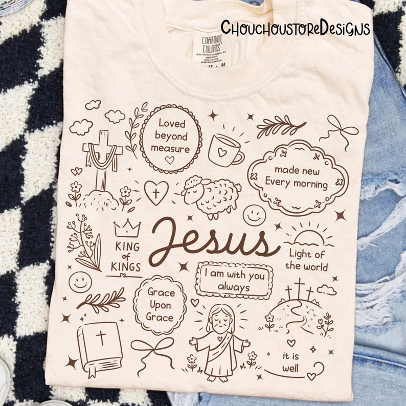 May include: Cream-colored t-shirt with a hand-drawn design featuring the word "Jesus" and various religious symbols and phrases, including a cross, heart, and "Light of the world". The text "CHOUCHOUSTORE DESIGNS" is at the top.