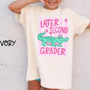 May include: Ivory-colored t-shirt with a pink and green graphic. The graphic features a cartoon alligator with a pink bow, the words "Later Second Grader", and pink stars. The shirt is a relaxed fit.
