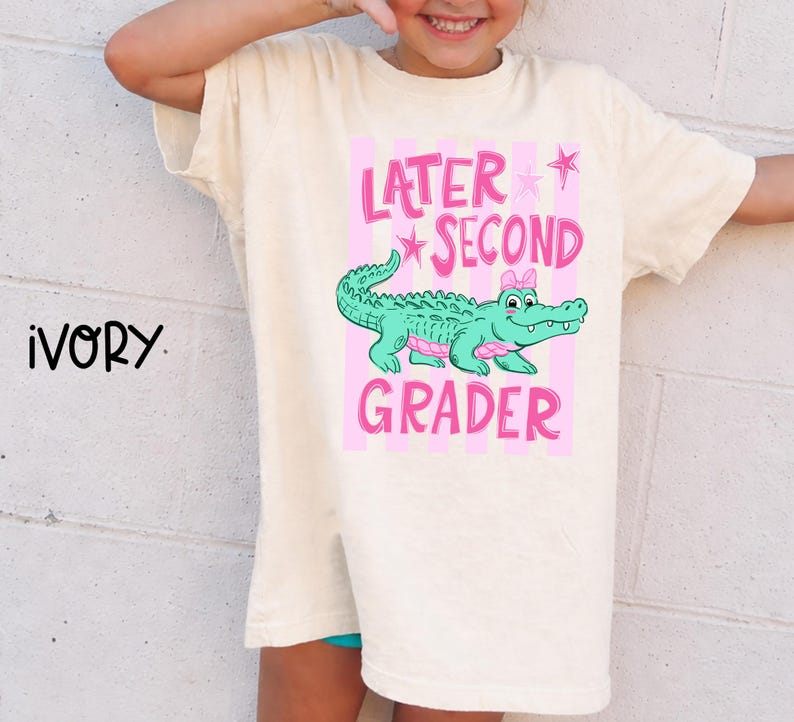 May include: Ivory-colored t-shirt with a pink and green graphic. The graphic features a cartoon alligator with a pink bow, the words "Later Second Grader", and pink stars. The shirt is a relaxed fit.