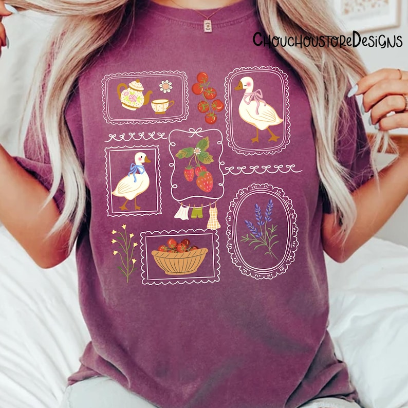 May include: A plum-coloured t-shirt featuring framed illustrations of a teapot, strawberries, a goose with a bow, and lavender. The shirt has the text "ChouchouStoreDesigns" printed on the upper right.
