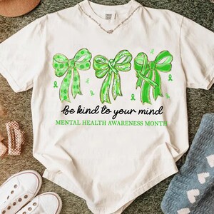 May include: Off-white t-shirt featuring three green bows with varying patterns and textures. The shirt includes the text "Be kind to your mind" and "Mental Health Awareness Month" in black script.