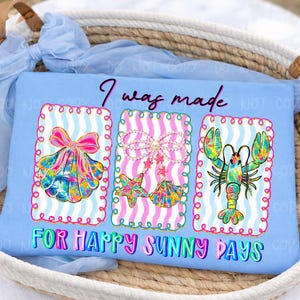 以下が含まれることがあります： 「I was made for happy sunny days」の文字が入った水色のTシャツ。 3つのフレームデザインがあり、リボン付きの貝殻、真珠付きのヒトデと貝殻、ロブスターが描かれています。 デザインは明るい色です。