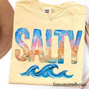 May include: Yellow Comfort Colors t-shirt with the word "SALTY" in colorful, textured lettering. Below the word are two blue ocean waves. The shirt has a small tag that says "COMFORT COLORS".