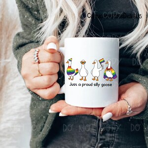 K&ouml;nnte beinhalten: Wei&szlig;e Keramik-Tasse mit vier Cartoon-G&auml;nsen mit Regenbogen-Akzenten und Accessoires. Die Tasse tr&auml;gt den Text "Just a proud silly goose" darunter. Die Tasse wird von einer Person gehalten.