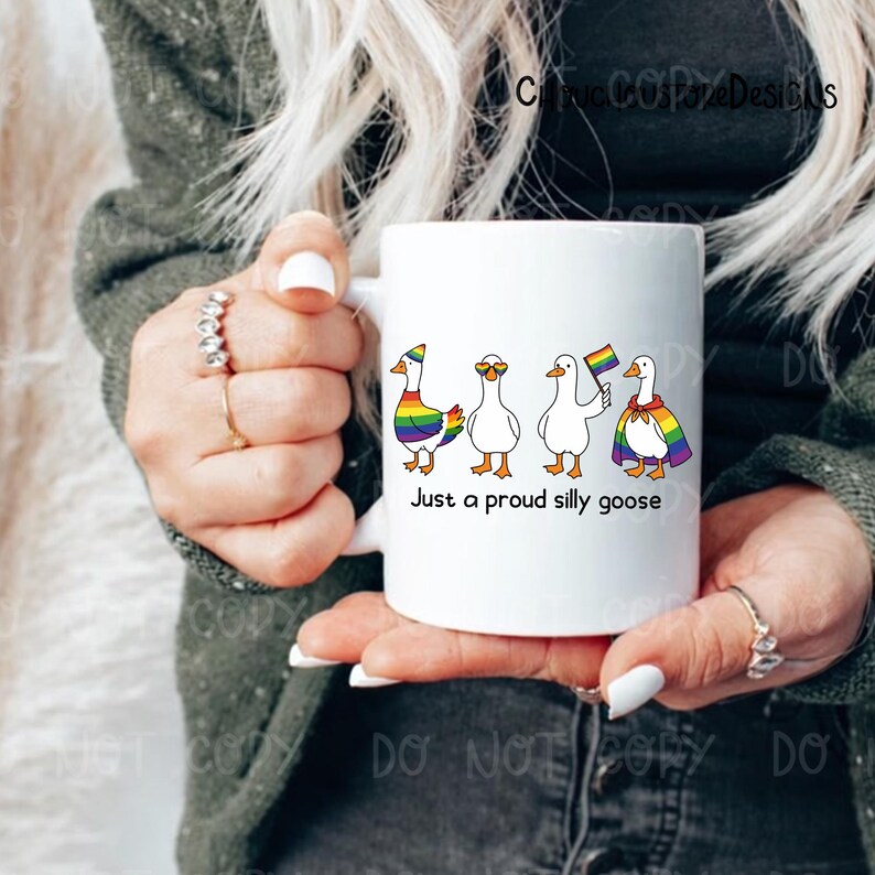 K&ouml;nnte beinhalten: Wei&szlig;e Keramik-Tasse mit vier Cartoon-G&auml;nsen mit Regenbogen-Akzenten und Accessoires. Die Tasse tr&auml;gt den Text "Just a proud silly goose" darunter. Die Tasse wird von einer Person gehalten.