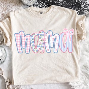 May include: Cream-colored t-shirt with the word "MAMA" in a patchwork design. The letters feature pink gingham, floral patterns, bow prints, and blue stripes. A pink bow is on the right side of the word.