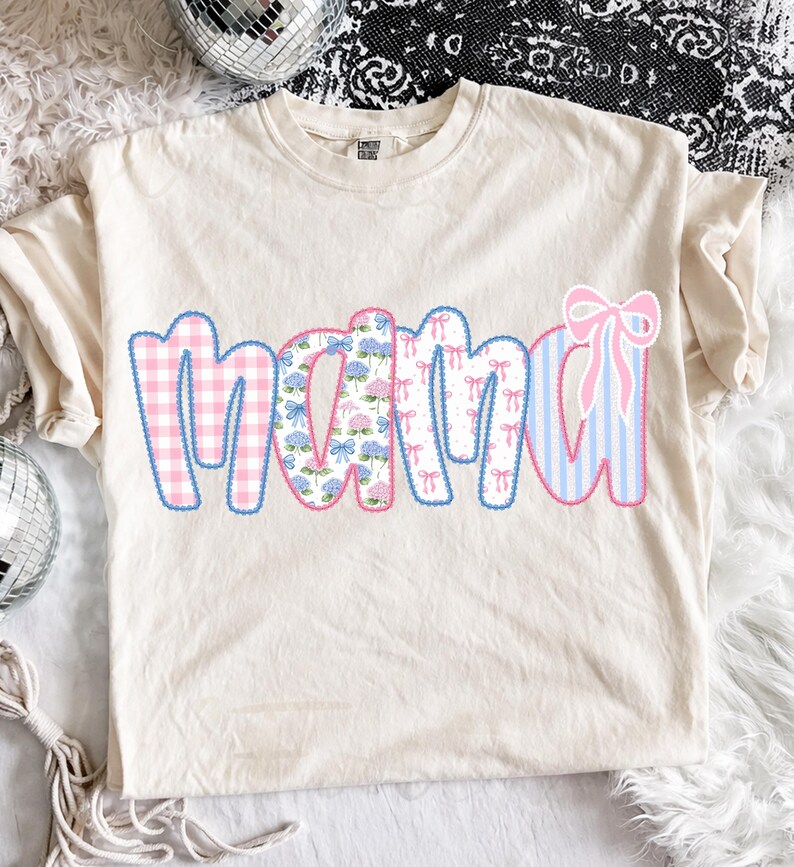 May include: Cream-colored t-shirt with the word "MAMA" in a patchwork design. The letters feature pink gingham, floral patterns, bow prints, and blue stripes. A pink bow is on the right side of the word.