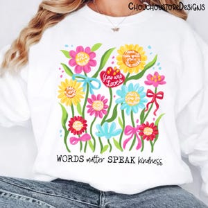 May include: White sweatshirt featuring a colorful floral design with inspirational quotes. The flowers are in shades of pink, yellow, and blue, with green leaves and bows. The text "WORDS matter SPEAK kindness" is at the bottom.