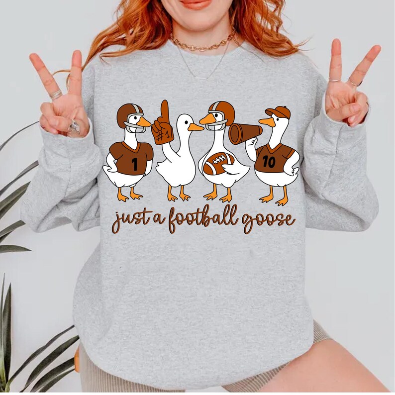 May include: A light gray sweatshirt featuring a graphic of four cartoon geese dressed in football gear. The geese wear helmets, jerseys, and hold a football and a megaphone. The text "just a football goose" is written below the geese.