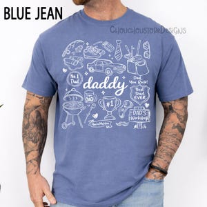 May include: Blue jean-colored t-shirt with a white graphic design featuring the word "daddy" and various icons like a car, grill, and trophy. The shirt also includes text that says "Dad You Rock!" and "Best Dad Ever".