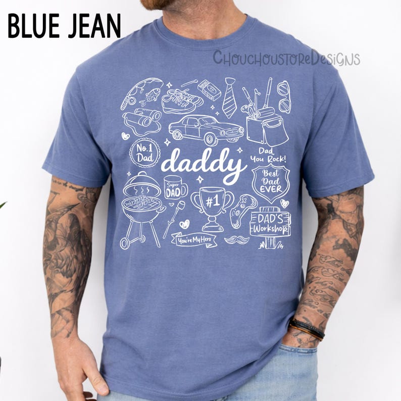 May include: Blue jean-colored t-shirt with a white graphic design featuring the word "daddy" and various icons like a car, grill, and trophy. The shirt also includes text that says "Dad You Rock!" and "Best Dad Ever".
