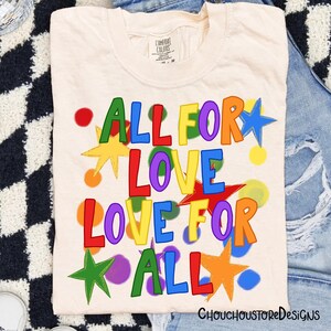 May include: Cream-colored t-shirt with the words "ALL FOR LOVE LOVE FOR ALL" in colorful, playful lettering. The design includes stars and polka dots in various colors. The shirt is folded on a denim surface.