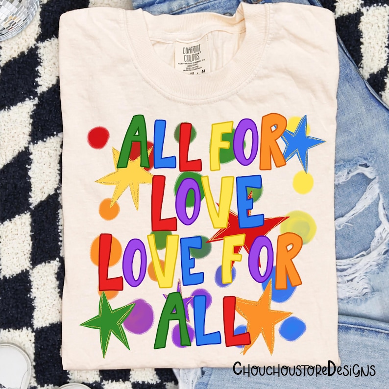 May include: Cream-colored t-shirt with the words "ALL FOR LOVE LOVE FOR ALL" in colorful, playful lettering. The design includes stars and polka dots in various colors. The shirt is folded on a denim surface.
