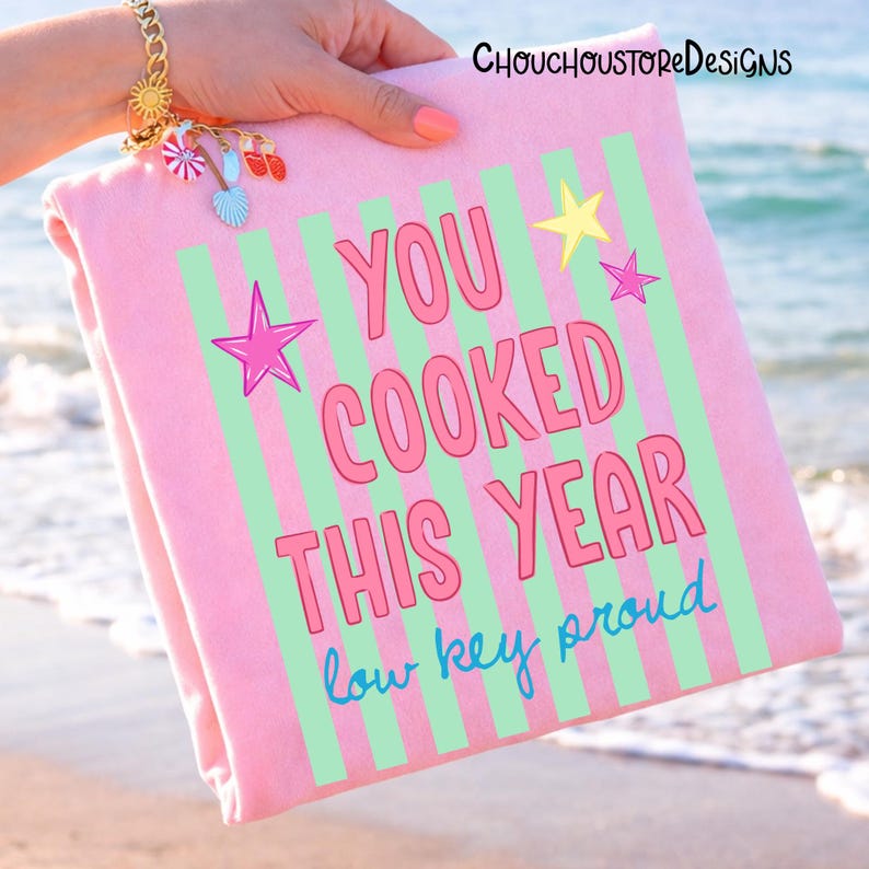 May include: A pink beach towel with green vertical stripes and the text "YOU COOKED THIS YEAR low key proud" in pink and blue. The towel is held up by a person with a gold bracelet with charms.