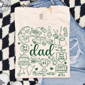 May include: Cream-colored t-shirt with a green graphic design featuring the word "dad" and various illustrations like a car, grill, and golf clubs. The shirt also includes text such as "No. 1 Dad" and "Best Dad Ever."