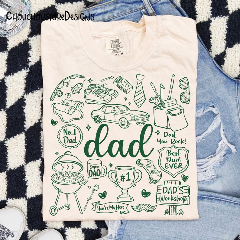 May include: Cream-colored t-shirt with a green graphic design featuring the word "dad" and various illustrations like a car, grill, and golf clubs. The shirt also includes text such as "No. 1 Dad" and "Best Dad Ever."