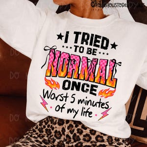 May include: White sweatshirt with the text "I TRIED TO BE NORMAL ONCE Worst 5 minutes of my life." The word "NORMAL" is in pink checkered letters with orange flames. Black stars and pink lightning bolts accent the text.