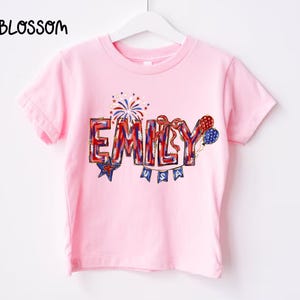 Puede incluir: Camiseta rosa claro con el nombre "EMILY" en letras rojas, blancas y azules. El dise&ntilde;o incluye fuegos artificiales, estrellas y globos, con "USA" debajo. La palabra "BLOSSOM" est&aacute; en la esquina superior izquierda.