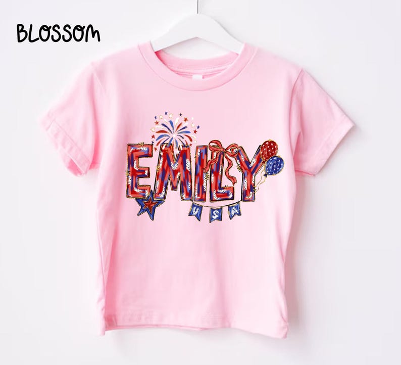 Puede incluir: Camiseta rosa claro con el nombre "EMILY" en letras rojas, blancas y azules. El dise&ntilde;o incluye fuegos artificiales, estrellas y globos, con "USA" debajo. La palabra "BLOSSOM" est&aacute; en la esquina superior izquierda.
