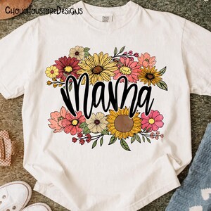 May include: White t-shirt featuring the word "Mama" in a black script font, surrounded by a colorful floral arrangement. The flowers include sunflowers, daisies, and other blooms in shades of yellow, pink, and red.