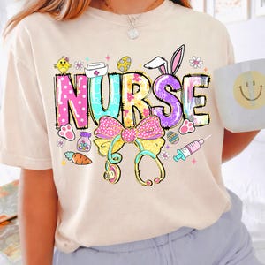 May include: A beige t-shirt featuring a colorful graphic design with the word "NURSE" in large, playful letters. The design includes Easter-themed elements like bunny ears, eggs, and a stethoscope, along with a bow and other medical symbols.