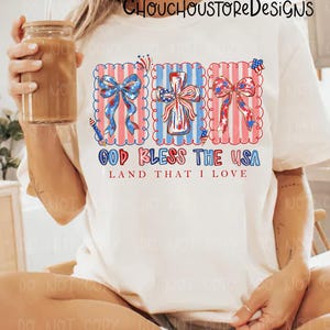 May include: Cream-colored t-shirt featuring a patriotic design with red, white, and blue bows, a cross, and the text "GOD BLESS THE USA LAND THAT I LOVE". The design is framed with a scalloped edge.
