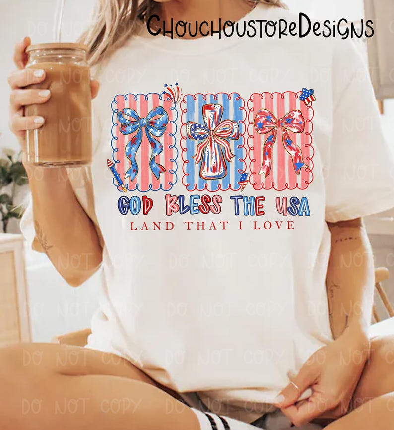 May include: Cream-colored t-shirt featuring a patriotic design with red, white, and blue bows, a cross, and the text "GOD BLESS THE USA LAND THAT I LOVE". The design is framed with a scalloped edge.