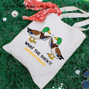 May include: A white canvas tote bag with a cartoon duck design. The ducks are brown, blue, and green, with yellow feet and beaks. Text on the bag reads "WHAT THE DUCK?! AWESOME QUACK!" The bag has long, cream-colored handles.