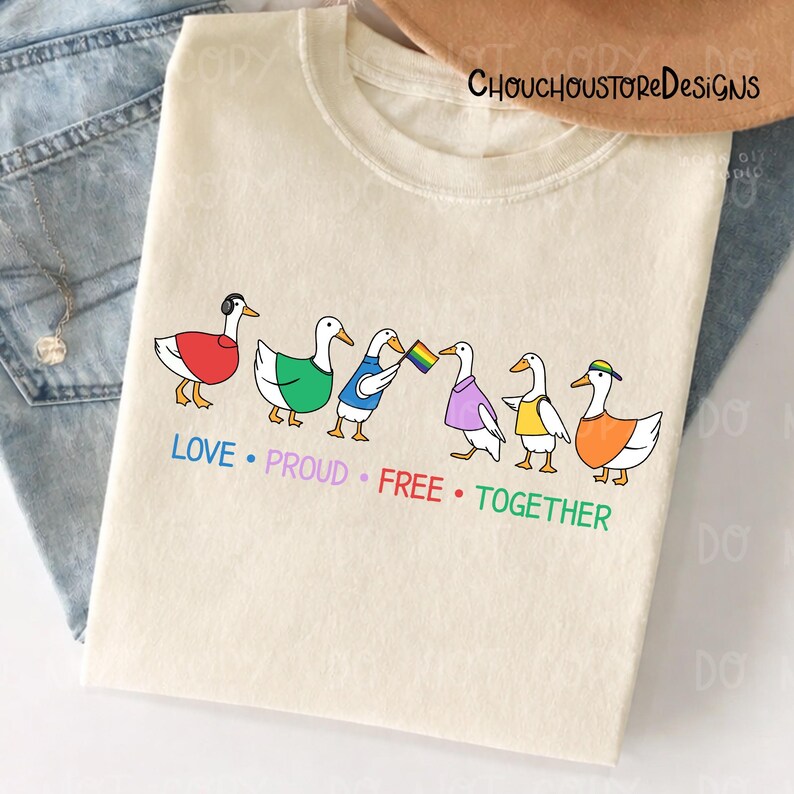 May include: Cream-colored t-shirt featuring a line of six cartoon ducks, each adorned with different colored accessories and holding a rainbow flag. The words "LOVE &bull; PROUD &bull; FREE &bull; TOGETHER" are printed below the ducks in a rainbow color scheme.