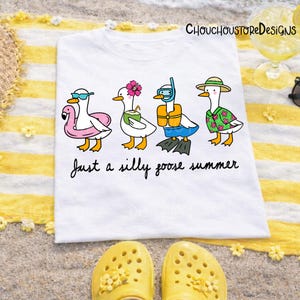 May include: White t-shirt with a whimsical illustration of four cartoon geese in summer attire. The geese are wearing sunglasses, a flower, a snorkel, and a Hawaiian shirt. The text "Just a silly goose summer" is below the geese.