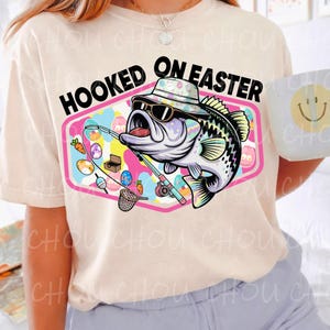 Pode incluir: Camiseta creme com a frase "HOOKED ON EASTER". O gr&aacute;fico apresenta um peixe de desenho animado usando &oacute;culos de sol e chap&eacute;u, segurando uma vara de pescar. Ovos de P&aacute;scoa e cenouras tamb&eacute;m est&atilde;o no design.