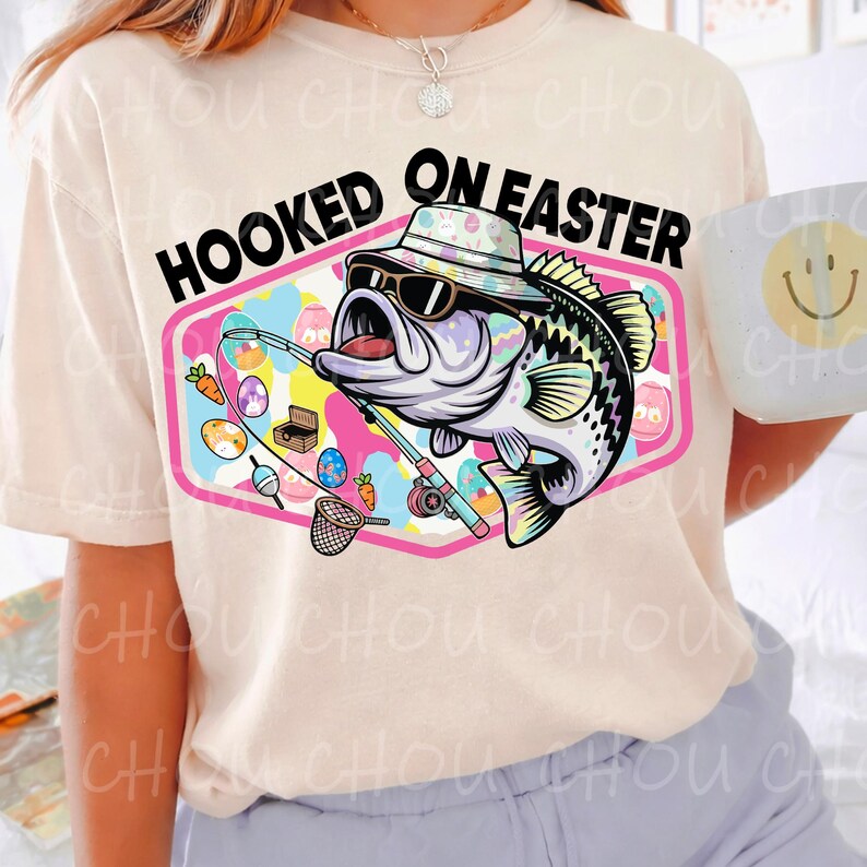 Pode incluir: Camiseta creme com a frase "HOOKED ON EASTER". O gr&aacute;fico apresenta um peixe de desenho animado usando &oacute;culos de sol e chap&eacute;u, segurando uma vara de pescar. Ovos de P&aacute;scoa e cenouras tamb&eacute;m est&atilde;o no design.