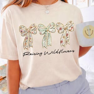 May include: A light beige t-shirt featuring three floral bow designs with gold outlines. The bows have a variety of floral patterns in pink, green, and yellow. The words "Raising Wildflowers" are written in a black script font below the bows.