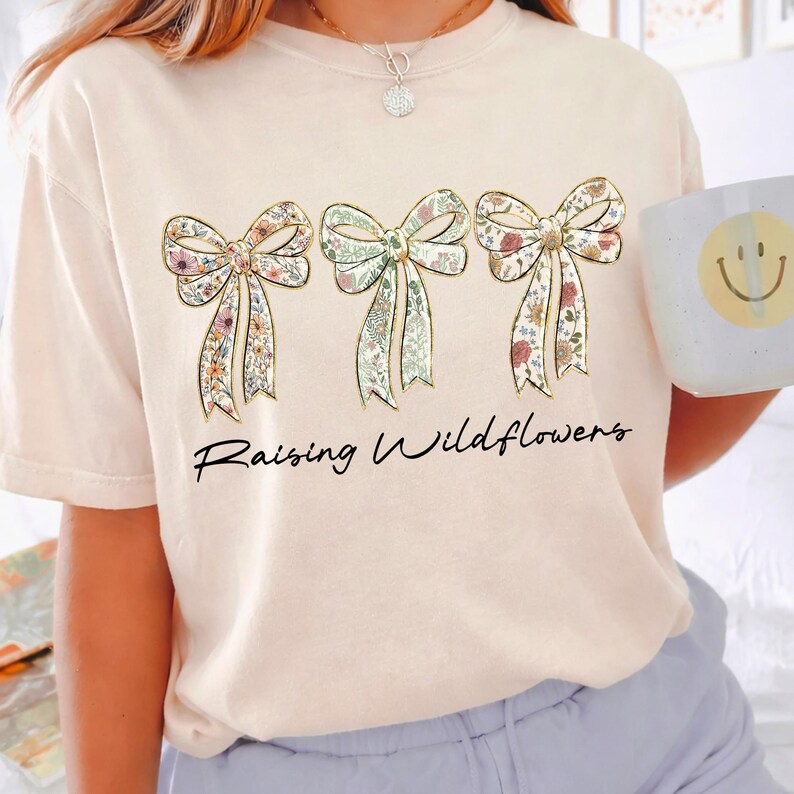 May include: A light beige t-shirt featuring three floral bow designs with gold outlines. The bows have a variety of floral patterns in pink, green, and yellow. The words "Raising Wildflowers" are written in a black script font below the bows.