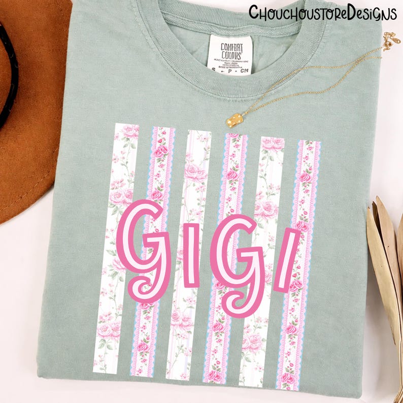 May include: Sage green t-shirt with the name "Gigi" in pink script. The shirt features vertical stripes with a floral pattern and lace trim. A gold necklace and a brown hat are visible.