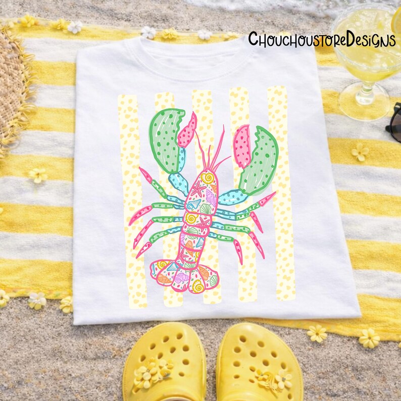 May include: White t-shirt featuring a colorful lobster design with green, pink, and blue accents. The lobster is decorated with seashells and starfish. The shirt is on a yellow and white striped towel with yellow sandals.