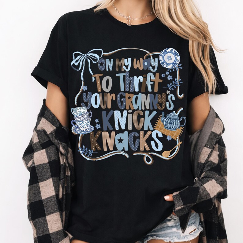 May include: Black t-shirt with the text "On my way to thrift your granny's knick knacks" in a whimsical font. The design includes illustrations of teacups, a teapot, and a decorative plate in blue and white.