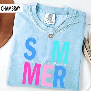 May include: Light blue t-shirt with the word "SUMMER" in blue, turquoise, and pink block letters. The word "vibes" is written in cursive script. A silver necklace with a pendant is draped on the shirt. The shirt is labeled "CHAMBRAY".