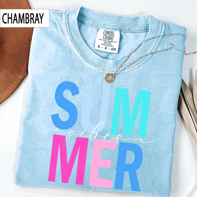 May include: Light blue t-shirt with the word "SUMMER" in blue, turquoise, and pink block letters. The word "vibes" is written in cursive script. A silver necklace with a pendant is draped on the shirt. The shirt is labeled "CHAMBRAY".