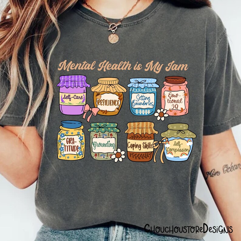 May include: Dark gray t-shirt with the text "Mental Health is My Jam." The shirt features colorful illustrations of jam jars labeled with mental health concepts like "Self-Care" and "Resilience."