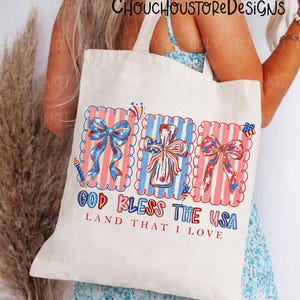 May include: A cream-colored tote bag featuring patriotic designs. The bag displays three framed images with red, white, and blue bows, a cross, and the text "GOD BLESS THE USA, LAND THAT I LOVE". The bag has a simple, rectangular shape.