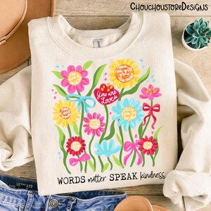 May include: Cream-colored sweatshirt with a floral design. The design features colorful flowers with inspirational phrases like "You are Loved" and "Words matter SPEAK kindness". A casual and uplifting garment.