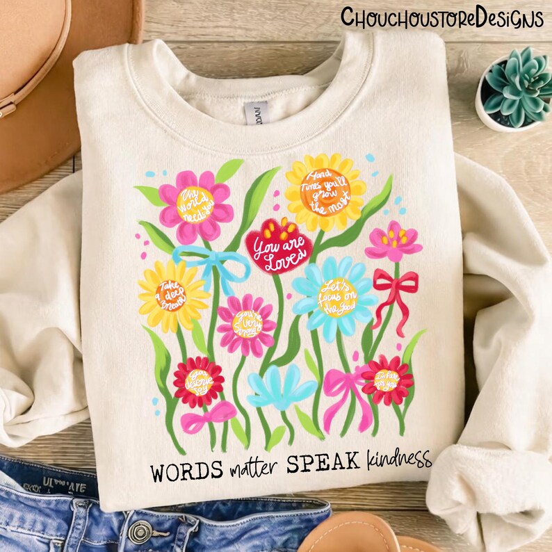 May include: Cream-colored sweatshirt with a floral design. The design features colorful flowers with inspirational phrases like "You are Loved" and "Words matter SPEAK kindness". A casual and uplifting garment.