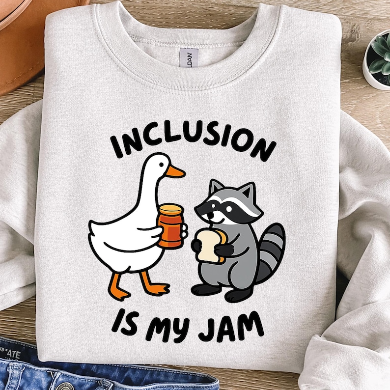 May include: Light gray sweatshirt with the words "INCLUSION IS MY JAM" in black. The graphic features a cartoon goose holding a jar of jam and a raccoon holding a slice of bread.