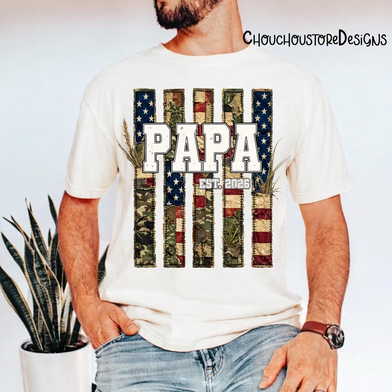 May include: Cream-colored t-shirt with a patriotic design. The design features the word "PAPA" in white block letters, with "EST. 2026" below. The background is a stylized American flag with camouflage and floral elements. The shirt is a casual wear item.