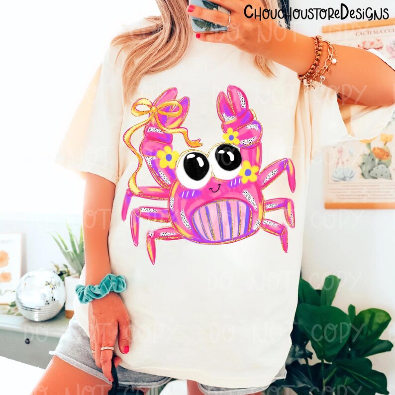 May include: Cream-colored t-shirt featuring a cartoon crab design. The crab is pink with yellow and purple accents, large black eyes, and a smiling face. The crab has a yellow bow and is decorated with yellow flowers. The text "ChouChouStoreDesigns" is at the top.